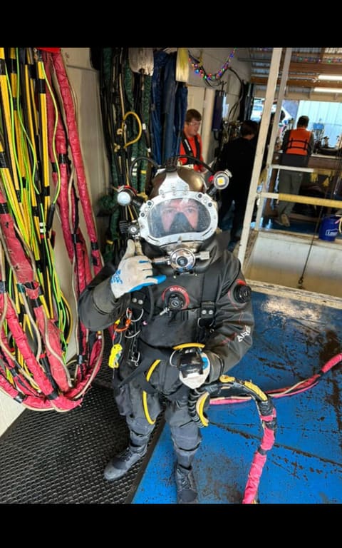 Commercial diver in a helmet and drysuit standing at the surface dive station with color-coded umbilicals hung behind