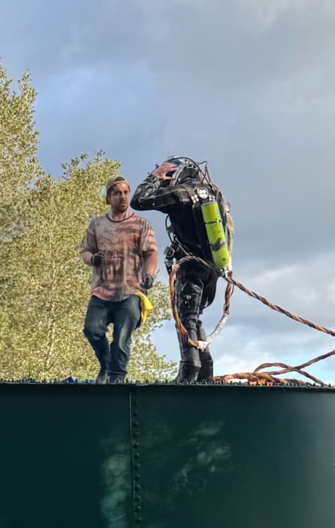 Commercial diver in a drysuit and helmet with a tender on top of a green bolted steel water storage tank, umbilical running to the hatch