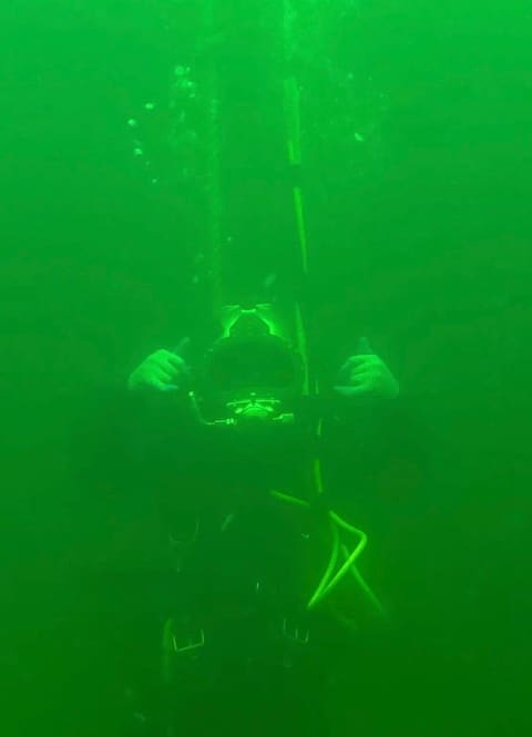 Diver descending a line inside a water storage tank, seen from below in clear green-lit water