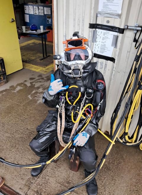 Diver seated in a black drysuit and dive helmet doing a pre-dive check with umbilical attached
